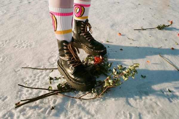 Black leather boots and colorful socks on snow with flowers, showcasing unique winter style.